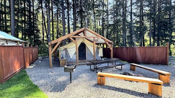 En trækonstruktion med saddeltag, omgivet af et træhegn, og med picnicborde og bænke.