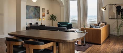 A modern living room with a wooden dining table, sofas, and a large window with curtains.