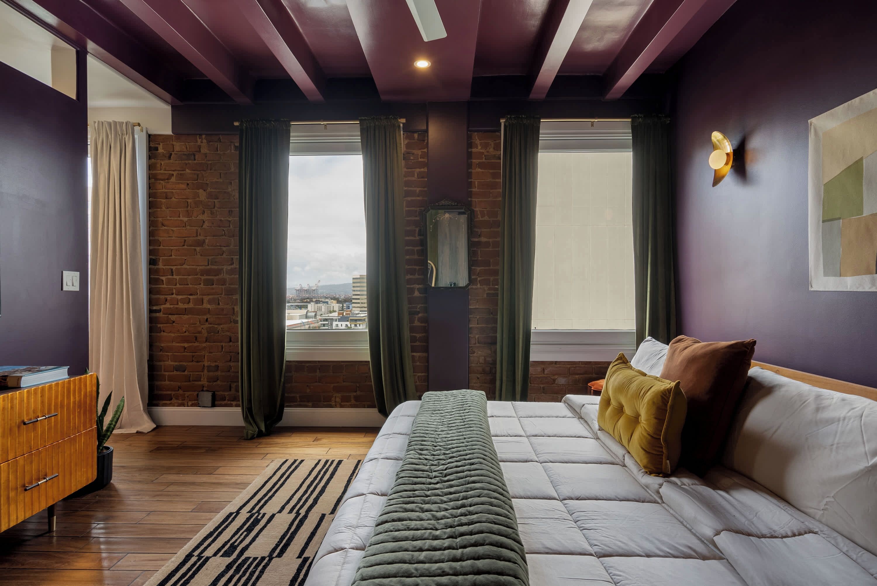 A modern bedroom with a brick wall, a large bed, a wooden dresser, and a view through the window.