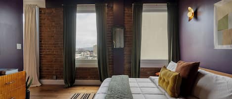 A modern bedroom with a brick wall, a large bed, a wooden dresser, and a view through the window.
