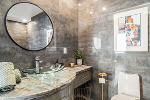 A bathroom with a glass bowl sink, a round mirror, a marble countertop, a toilet, and a framed abstract painting. - Ocean-View 1BR Condo in Historic Cooper Arms (Long Beach)