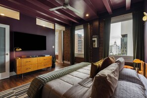 A modern bedroom with a large bed, a flat-screen TV, a wooden dresser, and a city view. - Ocean-View 1BR Condo in Historic Cooper Arms (Long Beach)