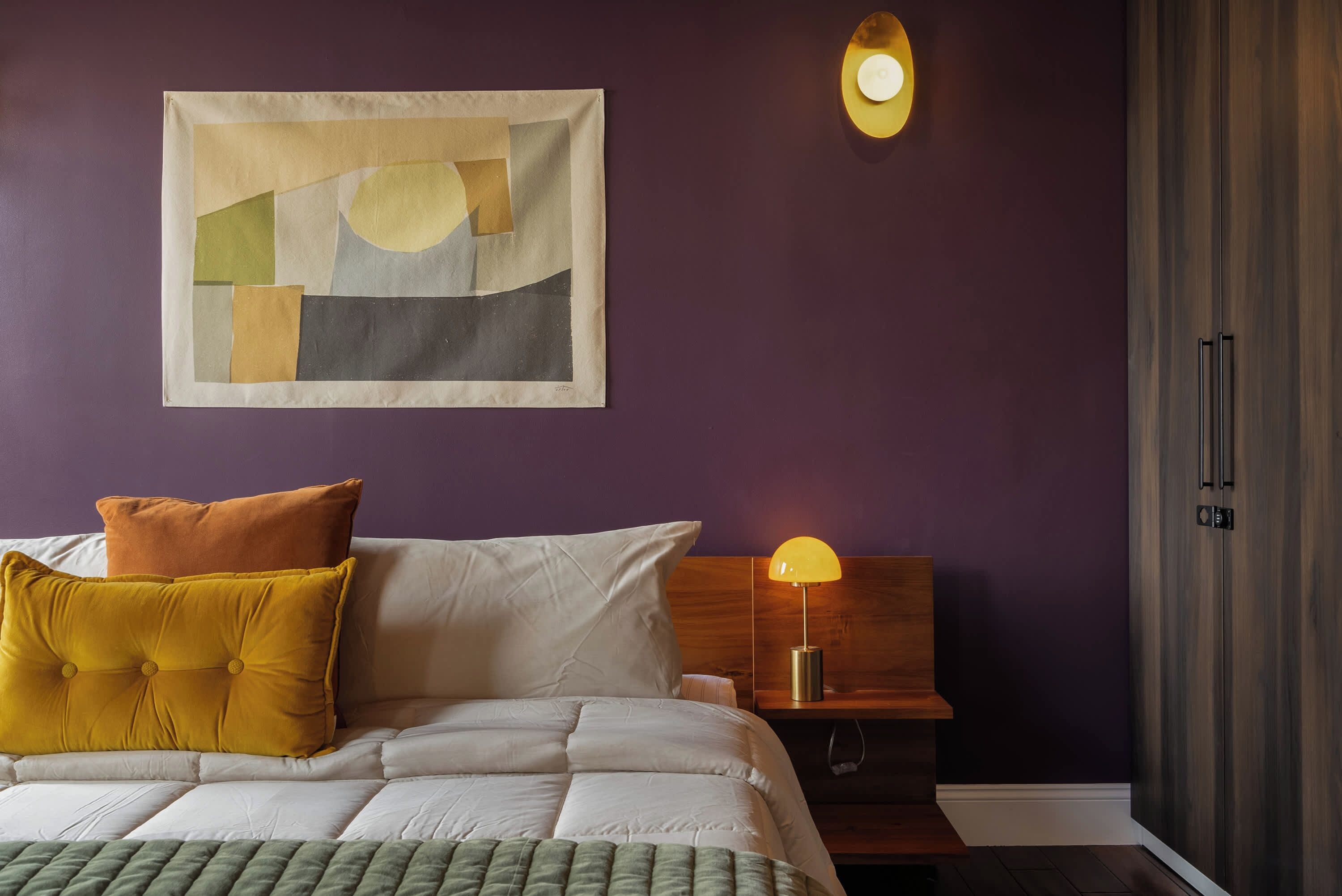 A neatly made bed with a green throw, a wooden nightstand with a lamp, and a wall-mounted abstract painting.