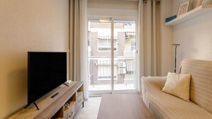 Una sala de estar con una televisión de pantalla plana sobre un mueble de madera, un sofá color beige y un ventanal corredizo que da a un balcón.