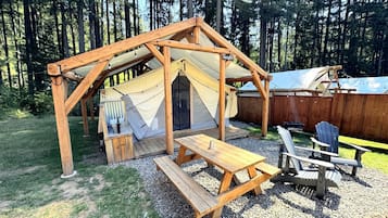 A wooden gazebo with a picnic table and Adirondack chairs, surrounded by a forest.