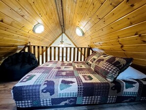 A loft bedroom with a wooden ceiling, a bed with a patterned quilt, and two ceiling lights.
