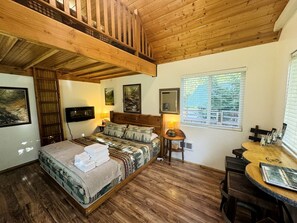A bedroom with a bed, a wooden ceiling, a window, and a small table with a lamp.