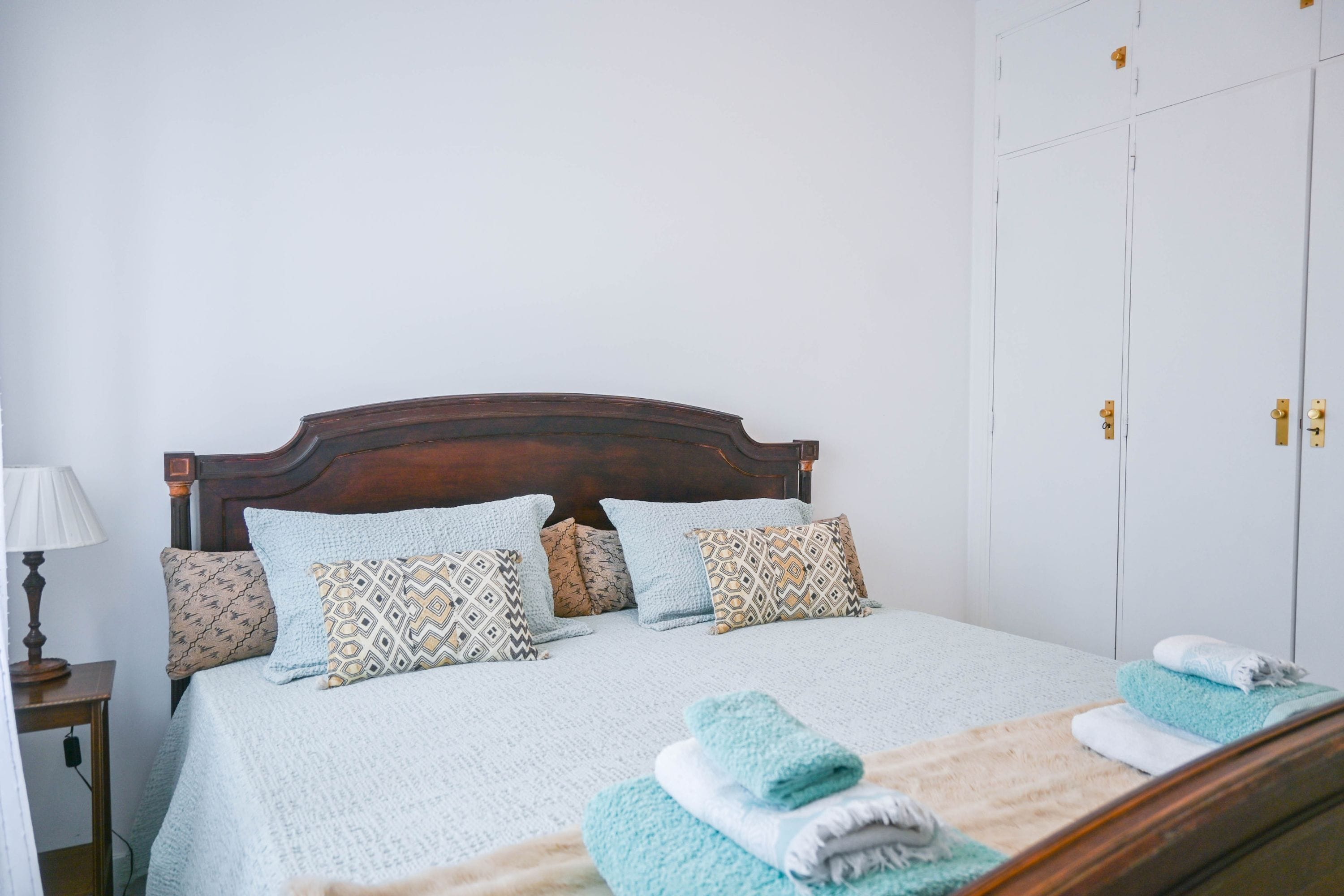 Una cama bien hecha con almohadas estampadas, una mesita de noche con una lámpara y un armario con espejo.