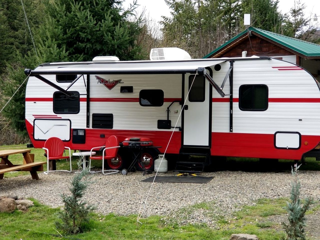 Ein Wohnmobil steht auf einem Campingplatz mit Picknicktisch und Stühlen, im Hintergrund ein kleines Gartenhaus und ein Kiesgrundstück.