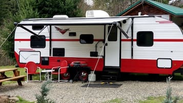 Ein Wohnmobil steht auf einem Campingplatz mit Picknicktisch und Stühlen, im Hintergrund ein kleines Gartenhaus und ein Kiesgrundstück.