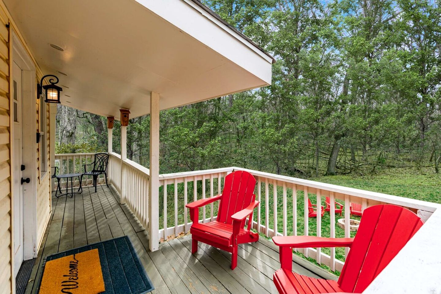 A wooden deck with a white railing, red Adirondack chairs, and a welcome mat.