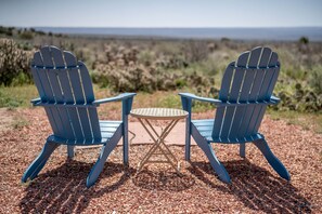 Two Adirondack chairs with a small round table in between, set on a gravel surface with a view of the ocean in the background.