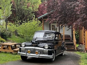 A vintage black car parked in front of a house with a wooden deck and a picnic table.