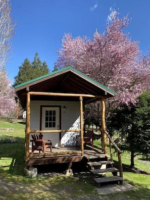 Una pequeña cabaña de madera con un porche, dos sillas y una escalera que lleva a la entrada.