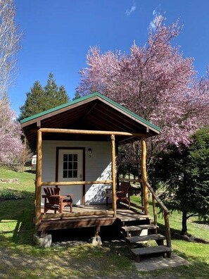 Una pequeña cabaña de madera con un porche, dos sillas y una escalera que lleva a la entrada.