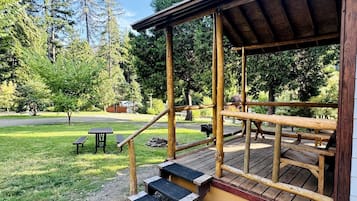 Una terraza de madera con escalones que conducen a una zona cubierta, rodeada de árboles y una mesa de picnic.