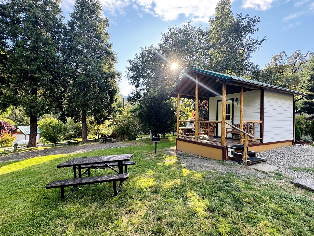 A small cabin with a porch, surrounded by trees and a picnic table.