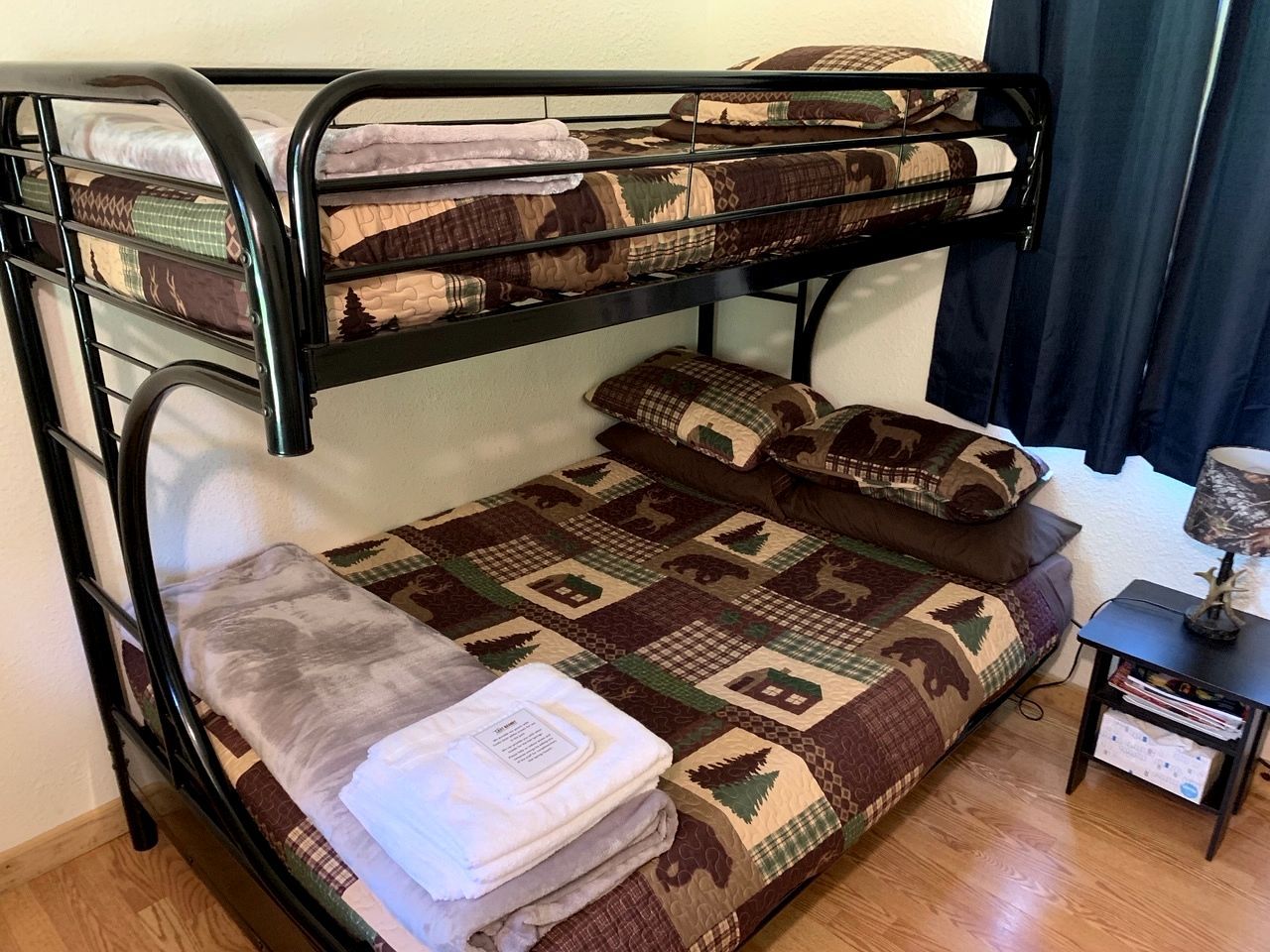 A bunk bed with a quilted bedspread and pillows, a small bedside table with a lamp, and a nightstand with books.