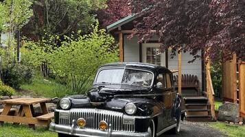Une voiture noire ancienne est garée devant une maison dotée d’une terrasse en bois et d’une table de pique-nique.
