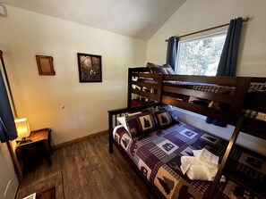 A bunk bed room with a wooden floor, a small bedside table with a lamp, and a window with curtains.