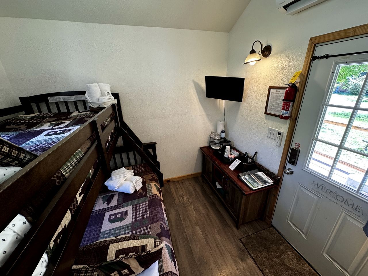 A room with a bed, a desk, a chair, a small table, a wall-mounted TV, a fire extinguisher, and a door with a welcome sign.