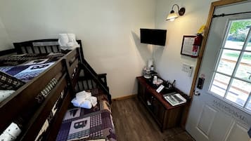 A room with a bed, a desk, a chair, a small table, a wall-mounted TV, a fire extinguisher, and a door with a welcome sign.