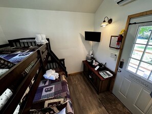Une chambre comprenant un lit, un bureau, une chaise, une petite table, un téléviseur fixé au mur, un extincteur et une porte sur laquelle est apposée une pancarte de bienvenue.