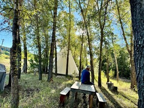 A person sitting at a wooden picnic table in a forested area with a tent in the background.