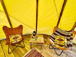 Interior de una carpa con dos sillas de madera, una mesita con libros y un farol, y una alfombra estampada.