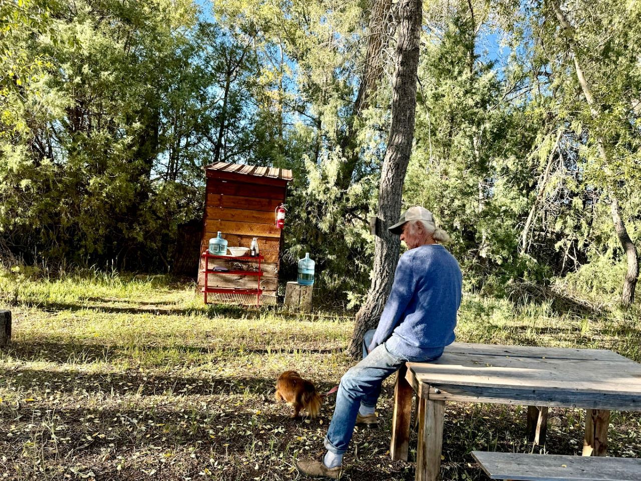 Um homem sentado em um banco numa área arborizada, com uma pequena construção de madeira ao fundo.
