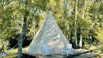 A teepee with two white chairs on a wooden platform, surrounded by trees.