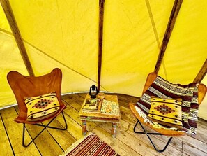A tent interior with two wooden chairs, a small table with books and a lantern, and a patterned rug.