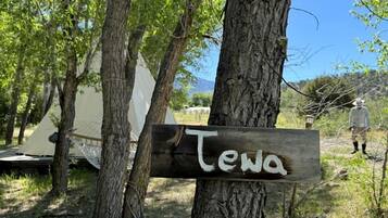 Ein hölzernes Schild mit dem Schriftzug 'Tewa', an einem Baum in einem Campingplatz mit Zelten und blauem Himmel.