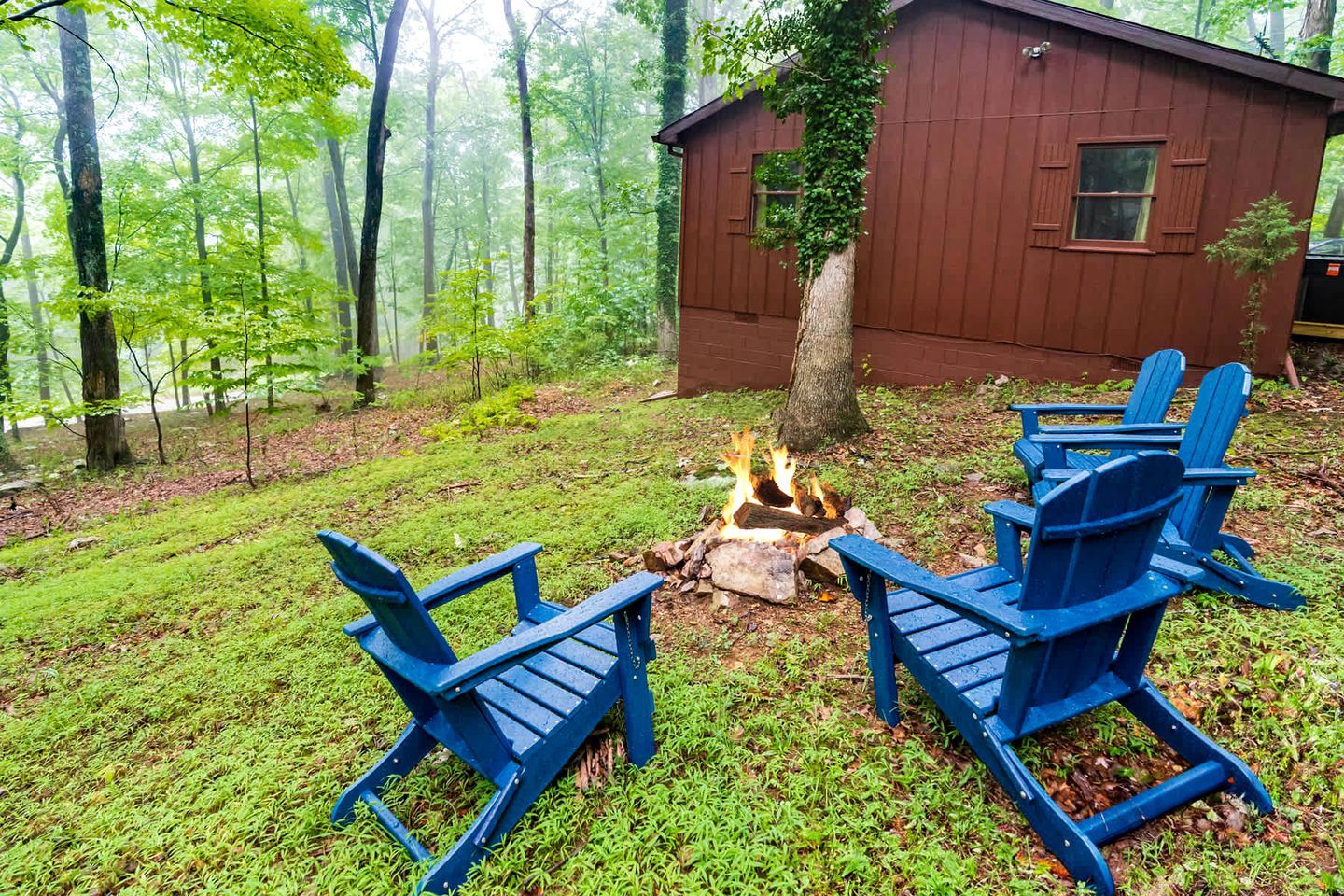 A cabin with a fire pit and blue Adirondack chairs in a forest setting.