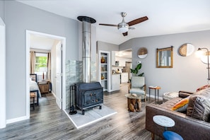 A modern living room with a fireplace, a ceiling fan, and a view into the kitchen. - Charming Dog-Friendly Cabin with Hot Tub and Fire Pit - Pond Access Makes This the Perfect Cabin for Your Fishing Getaway in Berkeley Springs, WV (Berkeley Springs)