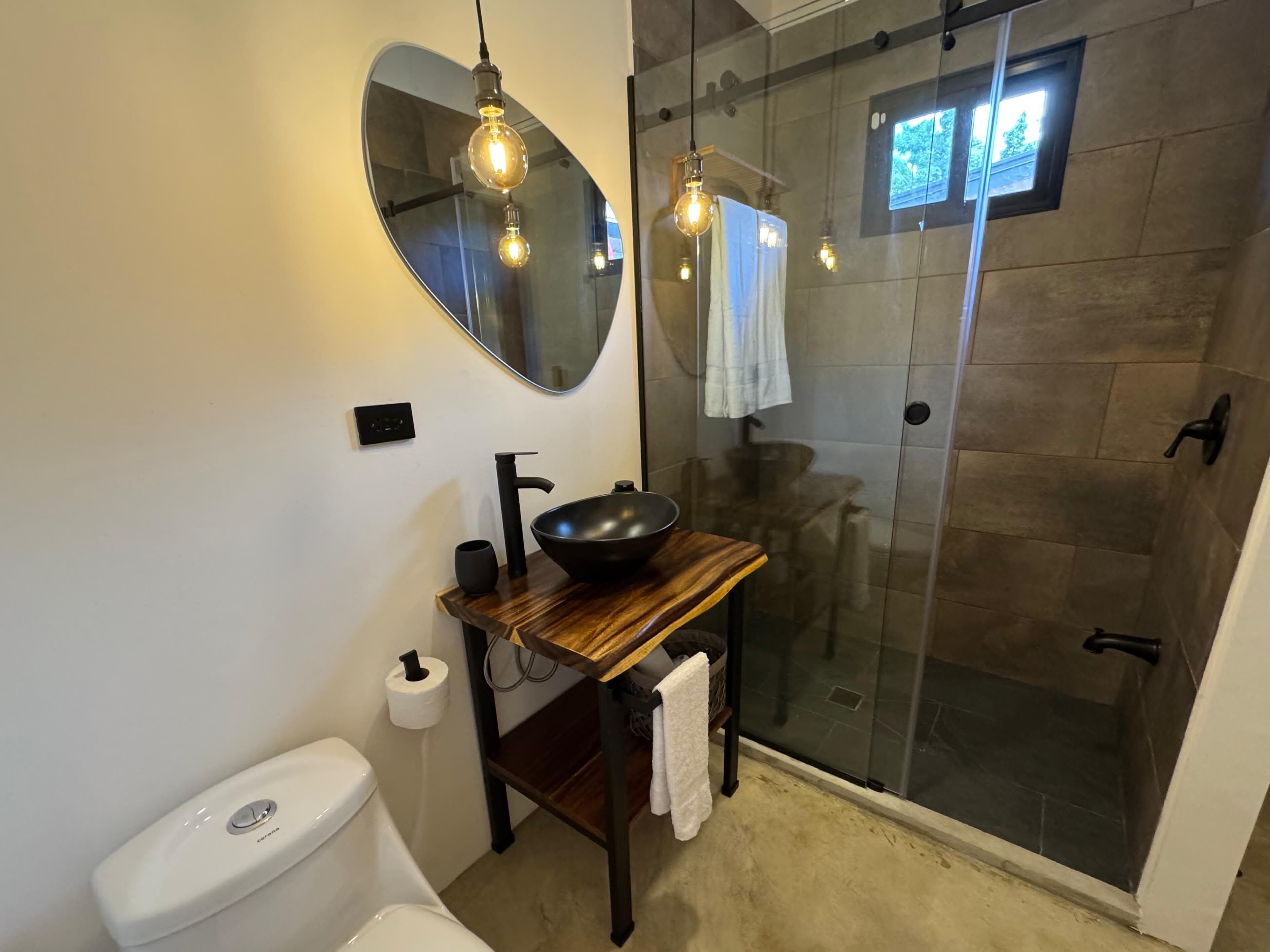Ein modernes Badezimmer mit rundem Spiegel, Waschbecken in Schwarz, Toilette und Duschkabine aus Glas.