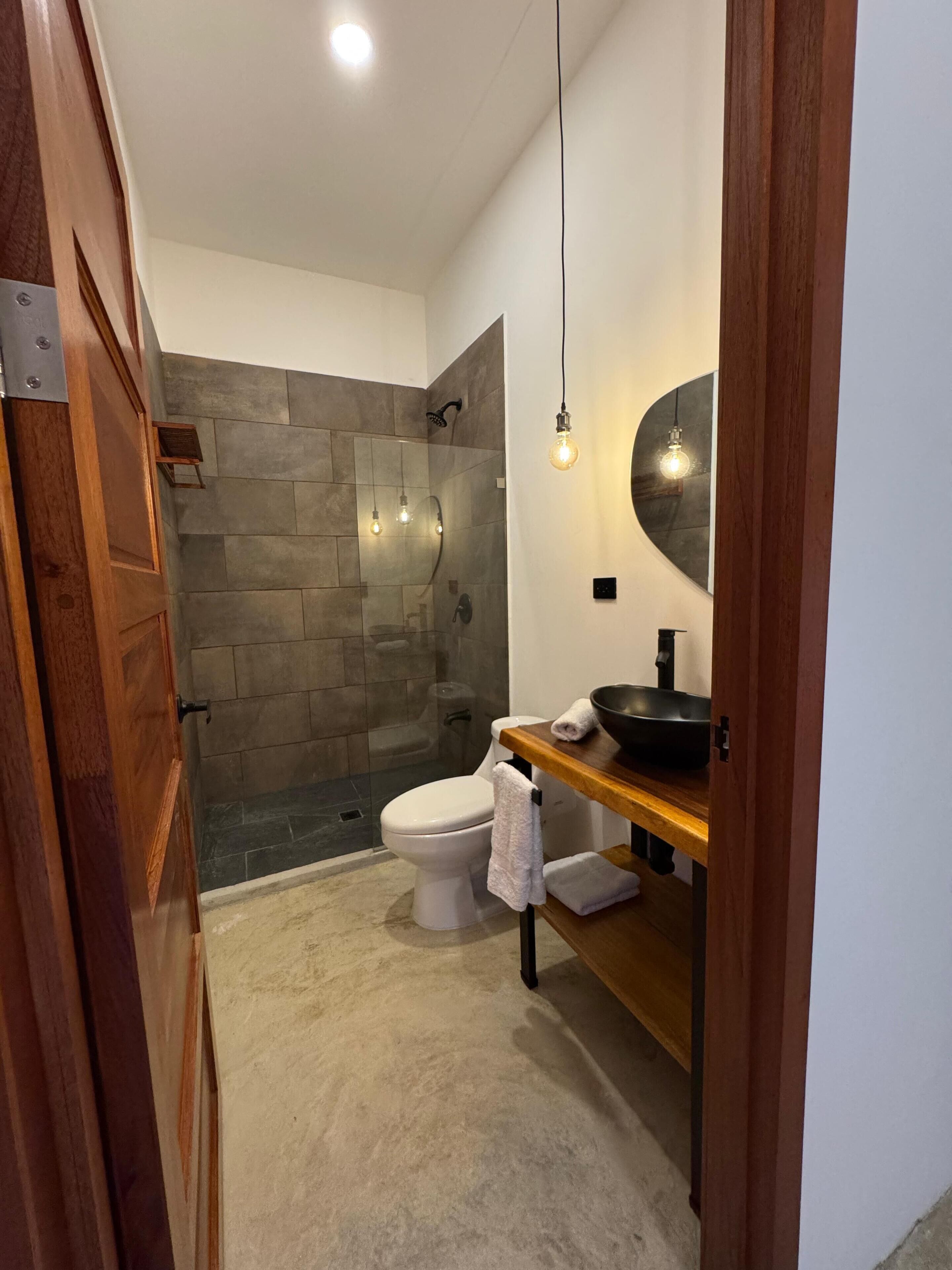 Une salle de bain moderne avec un mur de douche en pierre, des toilettes blanches et un meuble-lavabo en bois avec un évier noir.