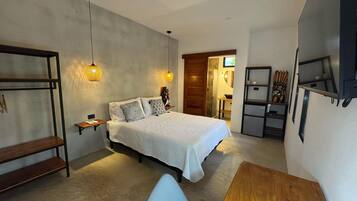A modern bedroom with a large bed, a wooden desk, and a small table. There is a shelf with decorative items and a door leading to another room.