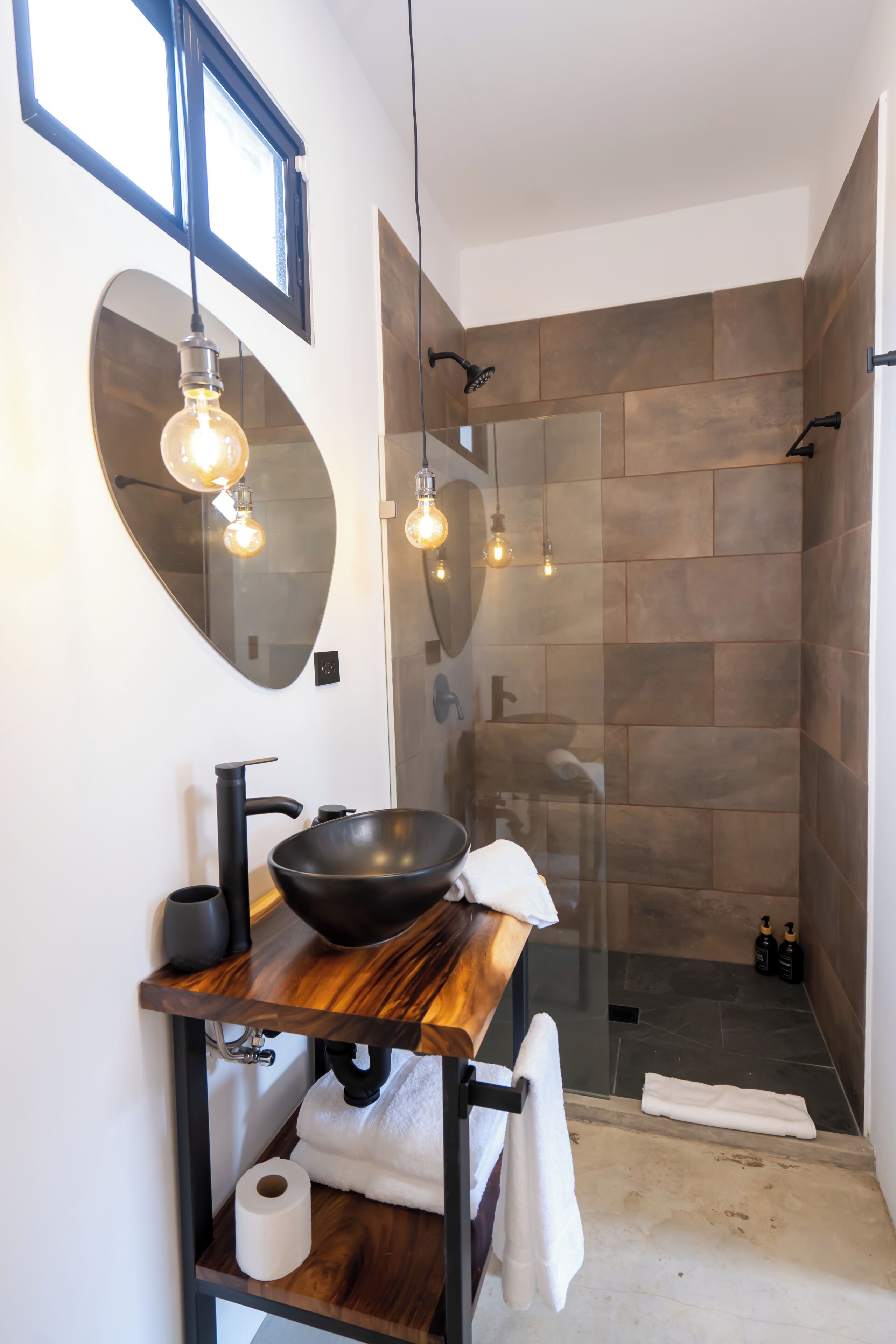 Un baño moderno con un espejo redondo, tocador de madera y ducha con cerramiento de vidrio.
