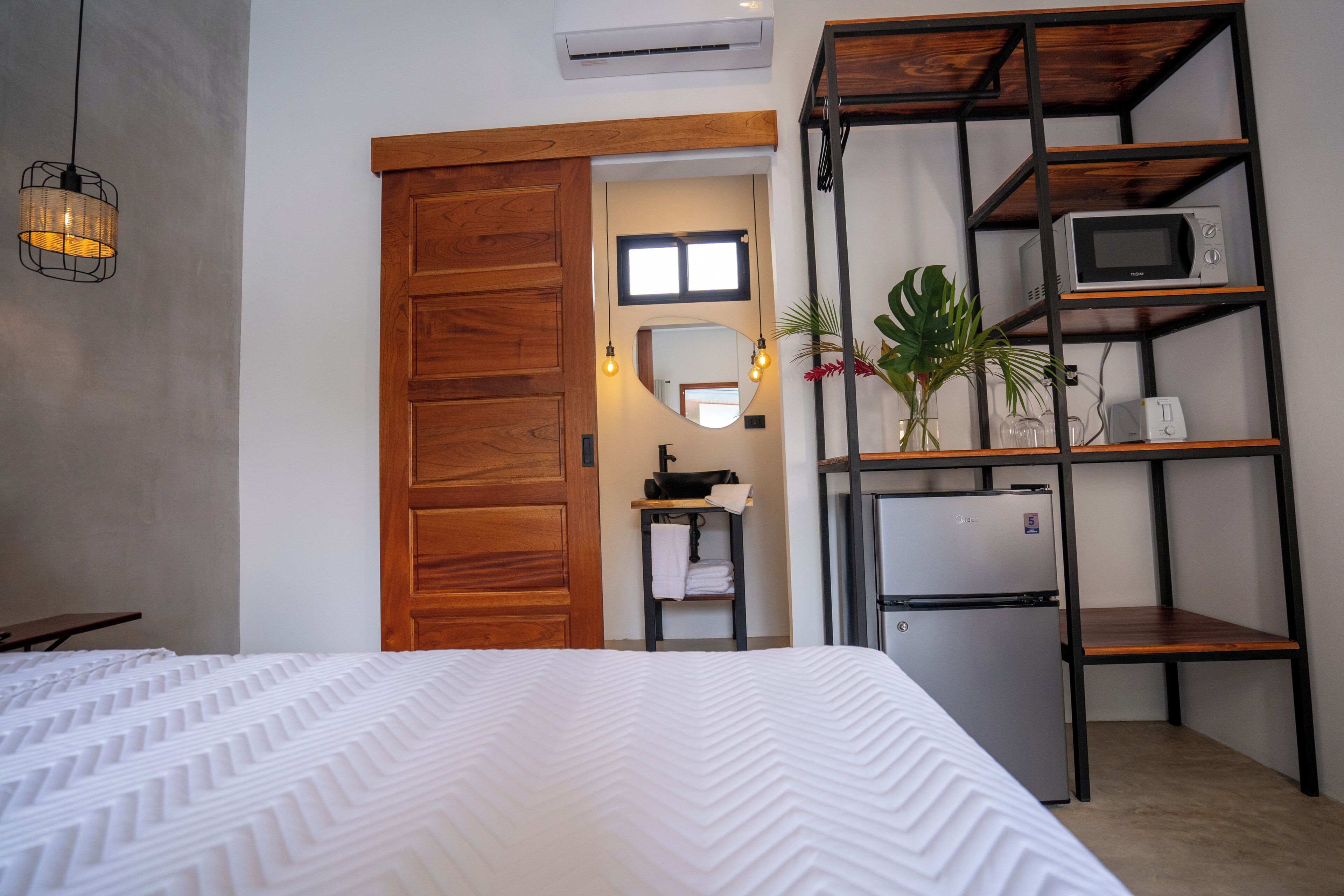 Ein Schlafzimmer mit einem Bett, einem Holzschrank, einem kleinen Tisch mit Handtuch, einem Regal mit Mikrowelle und einem Kühlschrank.