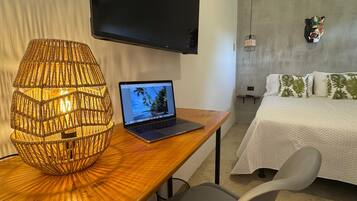 Et hotelværelse med et træskrivebord, en bærbar computer, en vævet lampe, en seng med hvide sengetøj og et ophængt tv.