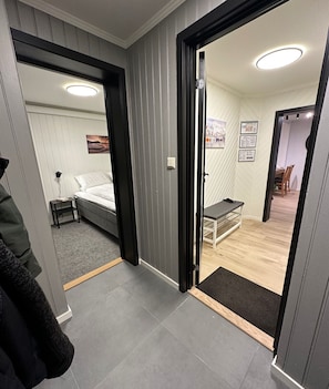 A modern hotel room with a bed, bedside table, and a small desk. There is a door leading to a dining area with a table and chairs.