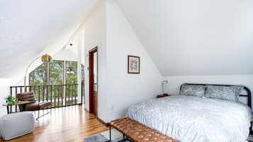A bedroom with a large bed, a bench, a desk, and a balcony with a view of trees.