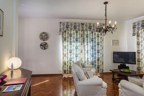 Ein Wohnzimmer mit einer Couch, einem Fernseher, einer Chandelierleuchtre und gemusterten Vorhängen.