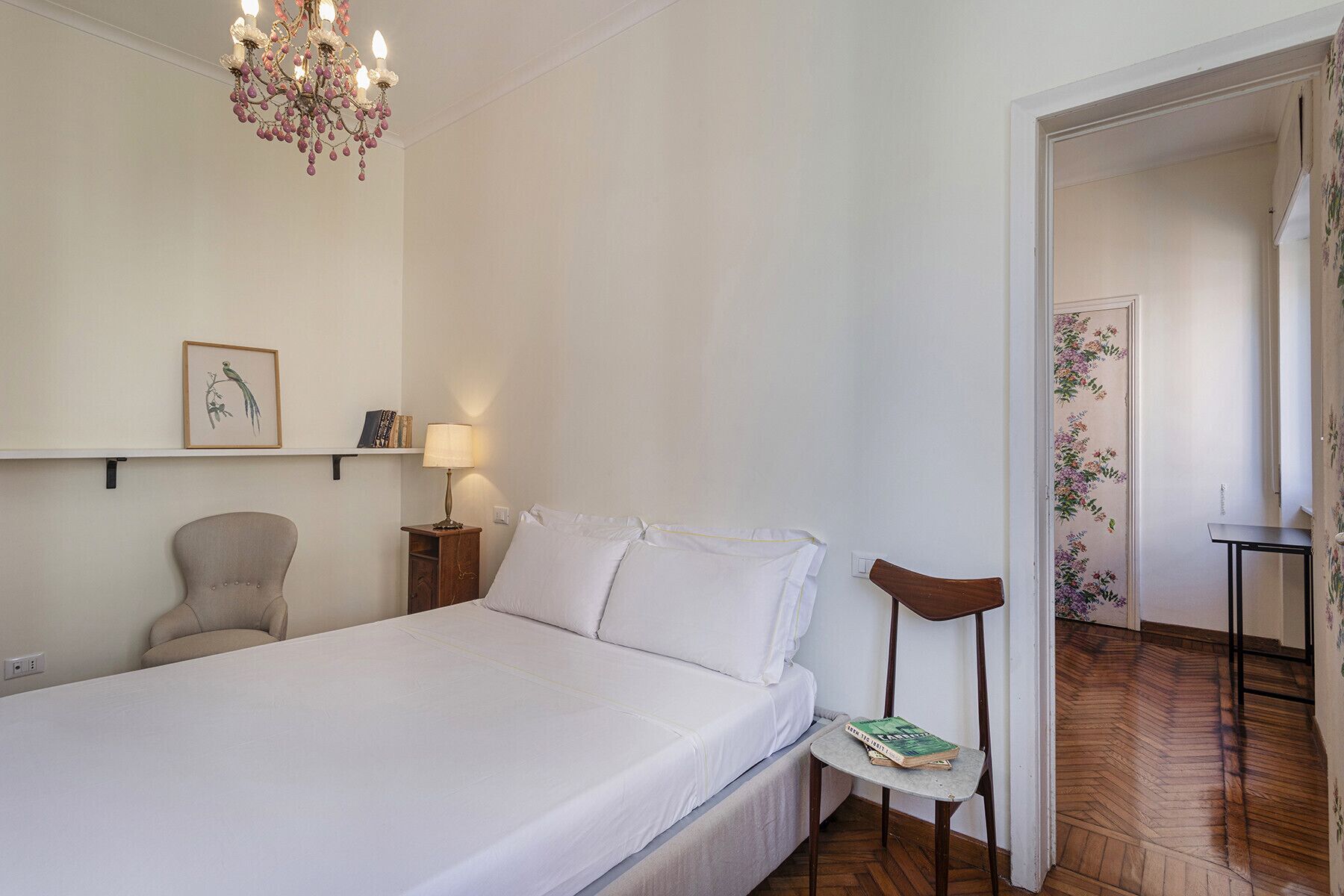 A bedroom with a bed, a chair, a small table with a book, a lamp, and a chandelier.