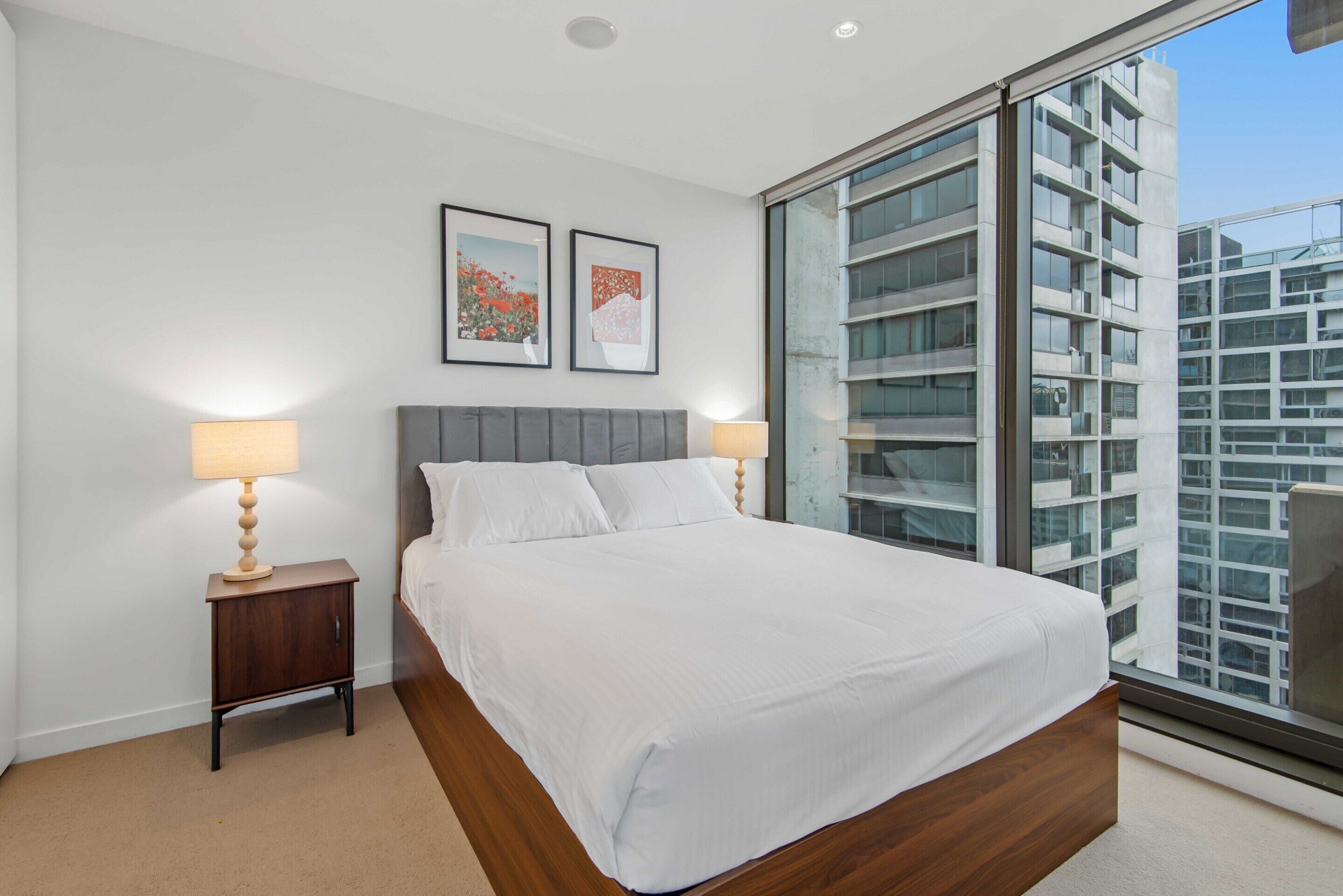 A modern bedroom with a large bed, two bedside tables, and a view of a cityscape through floor-to-ceiling windows.