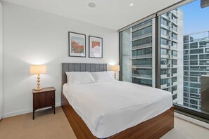 Ein modernes Schlafzimmer mit einem großen Bett, zwei Nachttischen und Blick auf eine Stadtlandschaft durch raumhohe Fenster.