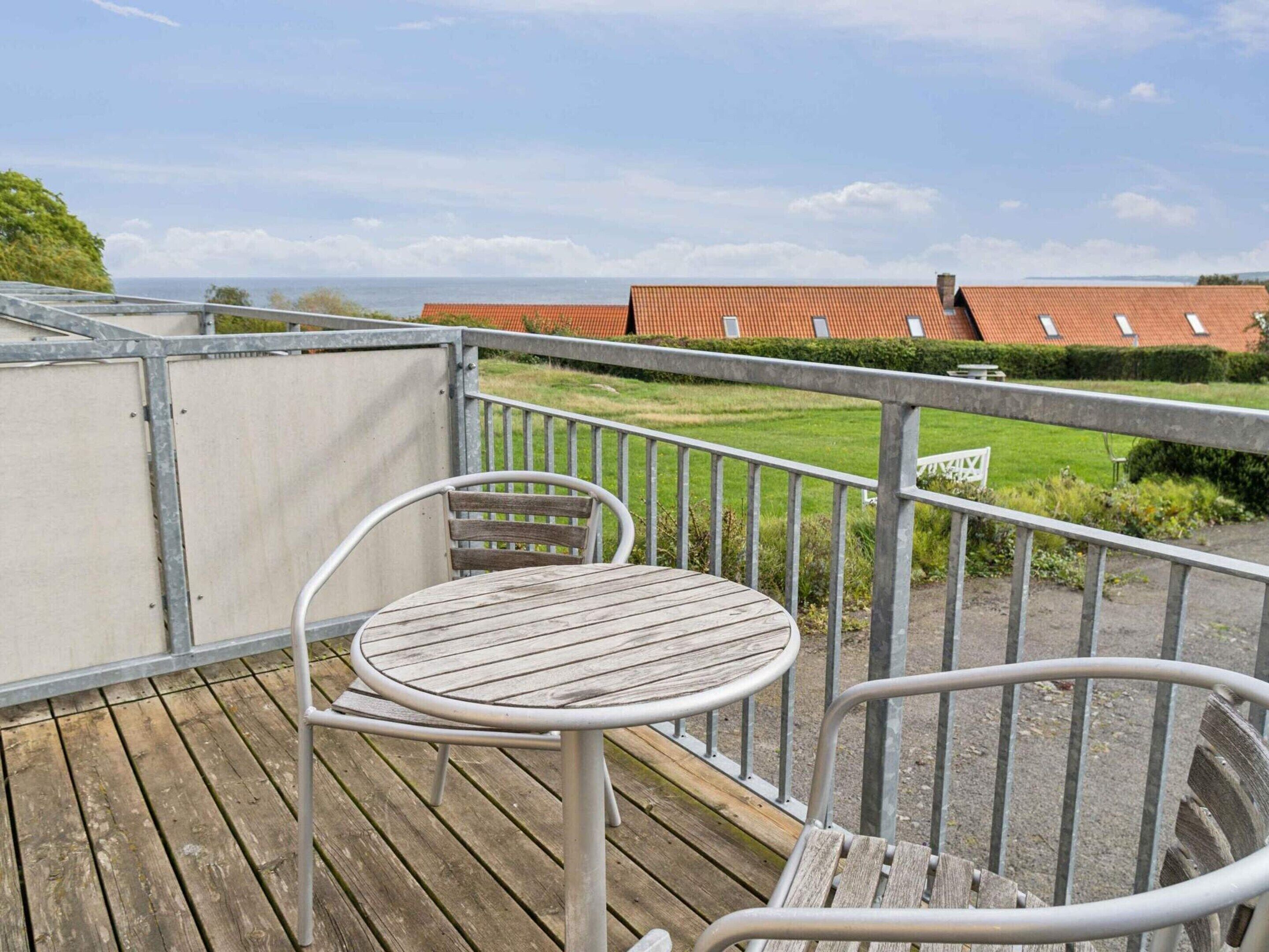 A balcony with a wooden table and metal chairs, overlooking a green lawn and a red-roofed building.