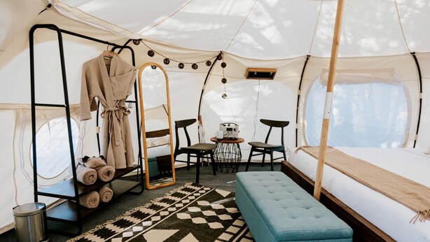 Een tent met een bed, een bank, een tafel met stoelen, een spiegel en een rek met handdoeken.