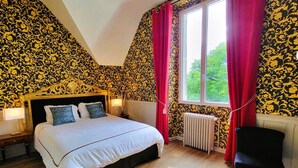 A bedroom with a large bed, a window with red curtains, and patterned wallpaper.